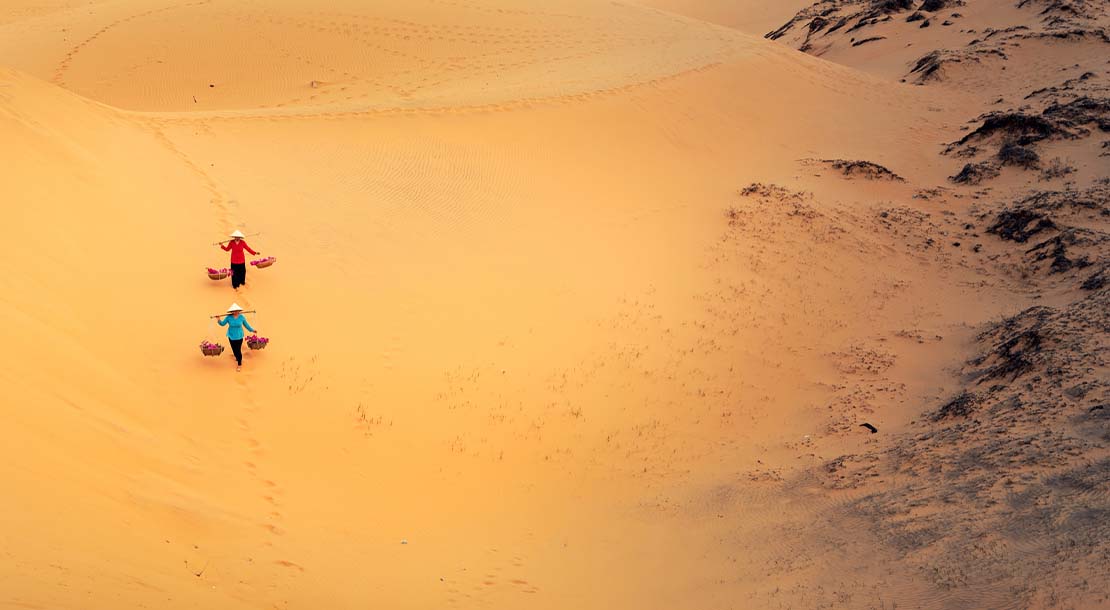 Two women carry baskets across a sand dune