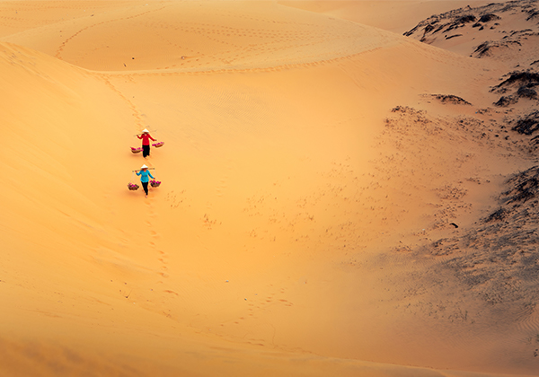 Two women carry baskets across a sand dune